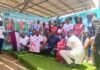 East End Lions Unveil New Jersey Ahead of FA Cup Final with Africell and Afrimoney Support Group of people on a stage posing for a photo at a sports event, wearing jerseys and formal attire with a colorful sponsor banner in the background.