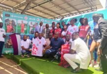 East End Lions Unveil New Jersey Ahead of FA Cup Final with Africell and Afrimoney Support Group of people on a stage posing for a photo at a sports event, wearing jerseys and formal attire with a colorful sponsor banner in the background.