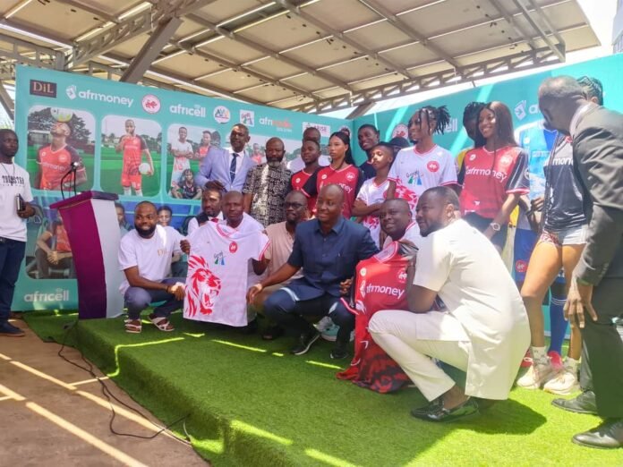 Group of people on a stage posing for a photo at a sports event, wearing jerseys and formal attire with a colorful sponsor banner in the background.