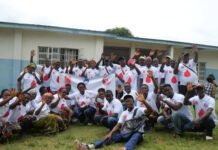 Lifeline Nehemiah, Ministry of Health Commemorate World Blood Donor Day with Voluntary Blood Donation Drive Large group of smiling people in white shirts with red logos posing outdoors, waving at the camera in front of a light blue building, with a banner in the center.
