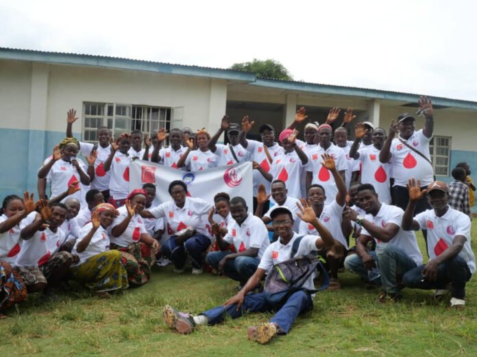 Large group of smiling people in white shirts with red logos posing outdoors, waving at the camera in front of a light blue building, with a banner in the center.