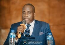 FCC, Police & Government Launch Multi-Agency Drive Against Illegal Dumping and Street Disorder Man in a dark blue pinstripe suit speaks into a handheld microphone at a formal event, with water bottles on the table and a government banner visible in the foreground.