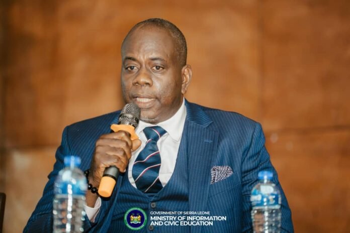 Man in a dark blue pinstripe suit speaks into a handheld microphone at a formal event, with water bottles on the table and a government banner visible in the foreground.