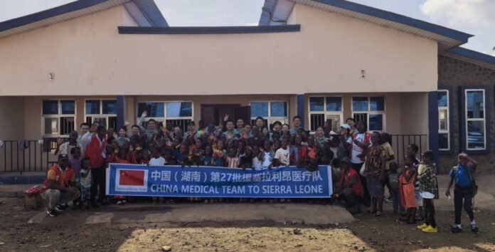 Group photo of a large medical team and local people in front of a building, holding a banner that reads 'China Medical Team to Sierra Leone'.