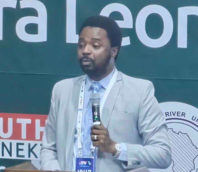 Man in a light gray suit speaks into a handheld microphone at a conference, with a large banner in the background.