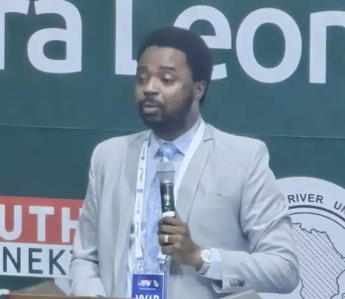 Man in a light gray suit speaks into a handheld microphone at a conference, with a large banner in the background.