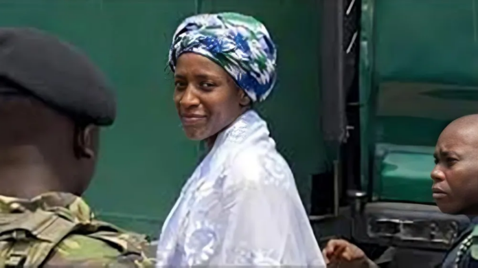 Zainab Sheriff Woman in a white patterned outfit and colorful headwrap smiles, flanked by two armed security personnel near a green vehicle.