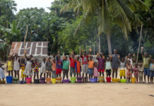 UNICEF Calls for Urgent Climate Action to Protect Sierra Leone’s Most Vulnerable Children Children stand in a line behind colorful buckets on a dirt ground, raising their hands amid a tropical village setting with palm trees.