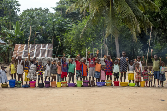 Children stand in a line behind colorful buckets on a dirt ground, raising their hands amid a tropical village setting with palm trees.