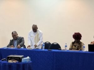 | The Calabash Newspaper Panel of four people seated at a long blue-cloth table; a man in white traditional attire stands to arrange papers while others look down at documents, with water bottles along the table.