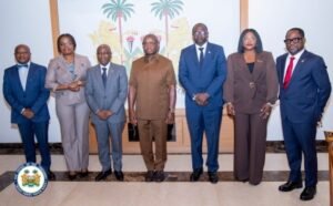 Group of seven formally dressed officials posing for a photo indoors, backdrop shows a palm-tree mural and an official seal at the floor.