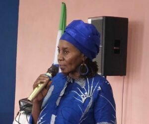 African woman in a blue traditional headwrap and matching outfit speaking into a microphone at an indoor event