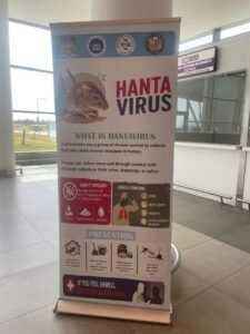 Roll-up banner about hantavirus with a rodent image and prevention tips, displayed in a public area near a tax/immigration desk.