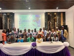Group of about 20 people posing for a conference photo behind a long table draped in purple and white, with a banner reading '2nd Conference' on a projection screen.