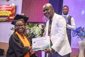 Graduate in cap and gown receives certificate from a man in a white blazer on stage with flowers behind them.