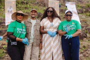 Group of four volunteers posing outdoors at an environmental event; two women in green 'Engage Change' shirts with blue gloves, a man in a khaki jacket, and a woman in a beige striped dress, all smiling. Behind them are banners promoting the cause.