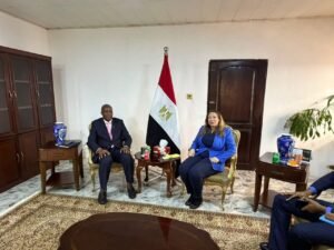 Two officials sit in an office: man in a dark suit with pink tie and woman in a blue blazer, flag of Egypt between them.