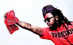 | The Calabash Newspaper Person with dreadlocks wearing a red shirt and sunglasses, arm extended to wave a red bandana.