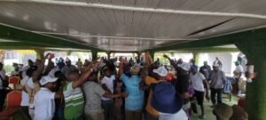 | The Calabash Newspaper Large group of people dancing and raising their arms under a shaded outdoor structure, suggesting a festive gathering or celebration.