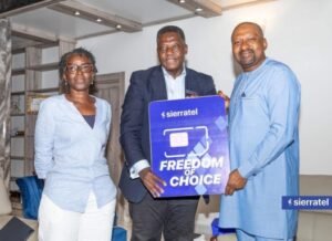 Three adults pose indoors, holding a blue Sierratel sign that reads 'FREEDOM OF CHOICE' during a presentation.