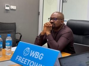 Vice Presidenta, Dr. Mohamed Juldeh Jalloh | The Calabash Newspaper Man in a maroon shirt with glasses sits at a conference table, hands clasped, in a meeting room; blue 'WBG FREETOWN' sign is on the table beside water bottles.