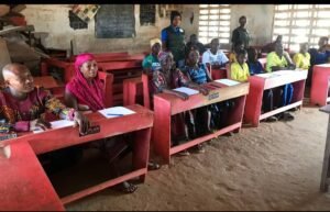 Students seated at red wooden desks in a classroom, writing on papers as a teacher watches from the back of the room.
