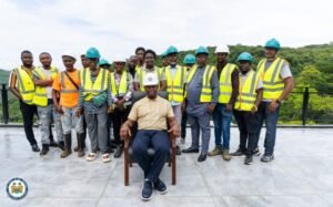 Group of construction workers in high‑visibility vests and hard hats standing behind a seated man in a tan shirt on a concrete rooftop with trees in the background.