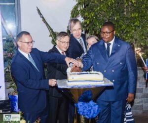 Five men in suits join hands over a rectangular cake on a pedestal, celebrating at a formal event.