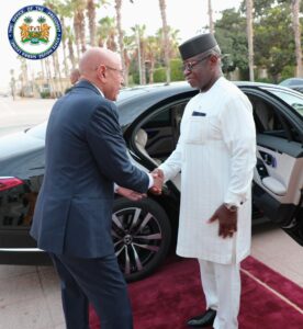 Two men greet each other with a handshake beside a black car; an official seal appears in the top-left corner, suggesting a formal event.