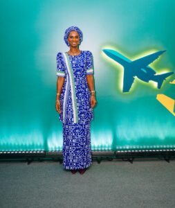 Full-body portrait of a woman in a blue and white patterned traditional dress and headwrap, standing in front of a teal backdrop with plane silhouettes on the right.