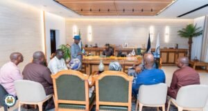 | The Calabash Newspaper Formal meeting in a government office: official sits at a large wooden desk with flags, addressing a group of people seated around the room.