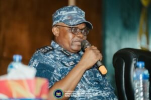 African man in blue camouflage uniform and cap, wearing glasses, speaking into a handheld microphone at a conference event.