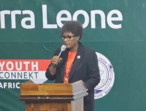 Speaker at a wooden podium delivering a talk, wearing glasses and an orange shirt, with a green banner behind.