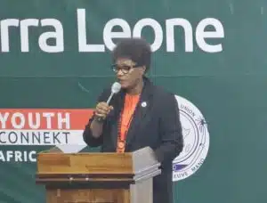 Speaker at a wooden podium delivering a talk, wearing glasses and an orange shirt, with a green banner behind.