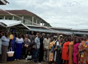 A large group of adults and children standing together outside a long, single‑story building with a metal roof, wearing colorful traditional and modern clothing.