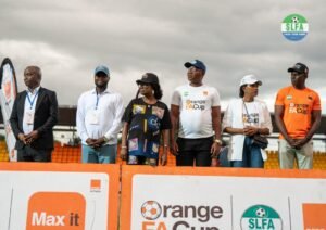 Six people stand on a sports event podium behind orange barriers with sponsor banners and logos visible (Orange FA Cup, SLFA) under a cloudy sky.