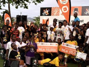 Group of children and teens at an outdoor awards event, posing with a trophy and large orange prize signs reading 'NLe 10,000' with banners in the background on a sports field