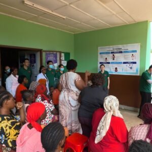 Group of people gathered in a hallway for a health outreach briefing, with workers in green shirts standing by informational posters on green walls while a woman speaks to the crowd.