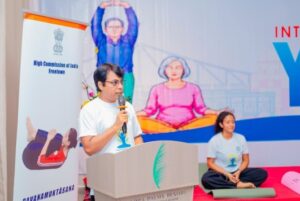 Man speaks into a microphone at a podium during a yoga event, with a banner for the High Commission of India in the background and a seated woman in a white shirt meditating nearby.