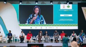 Female speaker on stage at a conference, displayed on a large screen, with a diverse panel seated behind her.