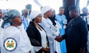 | The Calabash Newspaper Dignitaries at a formal event; a man in black shakes hands with a woman in white while others look on, with the Nigerian presidential seal visible in the foreground.
