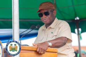 Man in a beige shirt and brown cap speaks at a wooden podium with a government seal visible, wearing sunglasses and a watch under a green canopy.