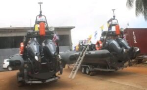 Two dark inflatable boats on trailers with large outboard motors, orange life jackets, and a metal ladder propped between them outside a warehouse.