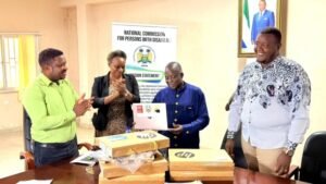 Four people stand around a table as a man in blue receives a document; others applaud in a room with a banner for the National Commission for Persons with Disabilities.