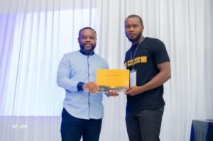 Two men exchange a certificate on stage during an award event; one in a light blue shirt hands it to a taller man in a black T‑shirt, with a gold certificate visible.