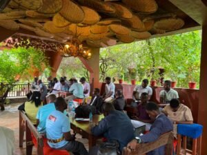 Group of people seated around a long table in an outdoor covered patio, working on laptops and papers.