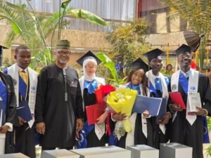 Group of graduates in caps and gowns posing for a photo at a ceremony, with one woman holding a bouquet and diploma while others display certificates; tropical plants and an indoor venue in the background.