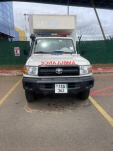 Front view of a white ambulance with red'AMBULANCE' lettering on the hood, blue and red emergency lights, Toyota emblem, and license plate AXG 163.