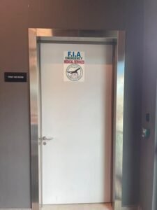 White door in a stainless steel frame with an'FIA Emergency Medical Services' sign and emblem; a wall label to the left reads 'First Aid Room'.