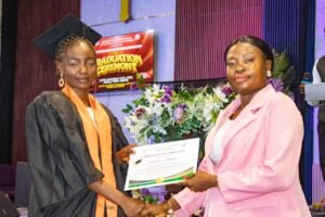 | The Calabash Newspaper Graduate in cap and gown receives a certificate from a woman in a pink blazer at a graduation ceremony with a floral backdrop.